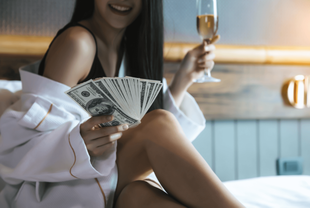 A joyful woman in a white robe holding a stack of hundred-dollar bills and a glass of champagne, symbolizing financial success and freedom.